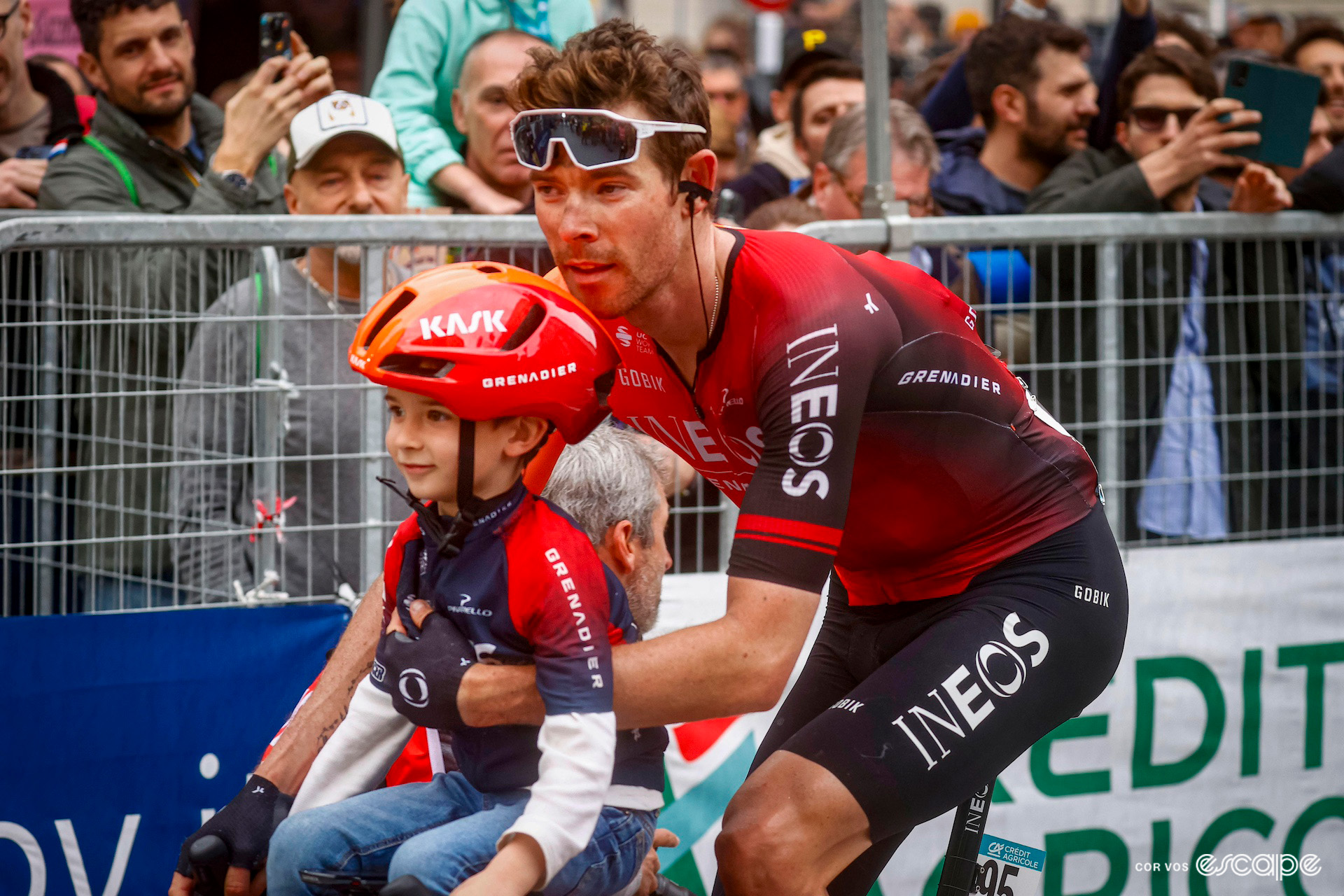 Luke Rowe with his young son perched on his handlebars in his helmet after the finish of Milan-San Remo 2024.
