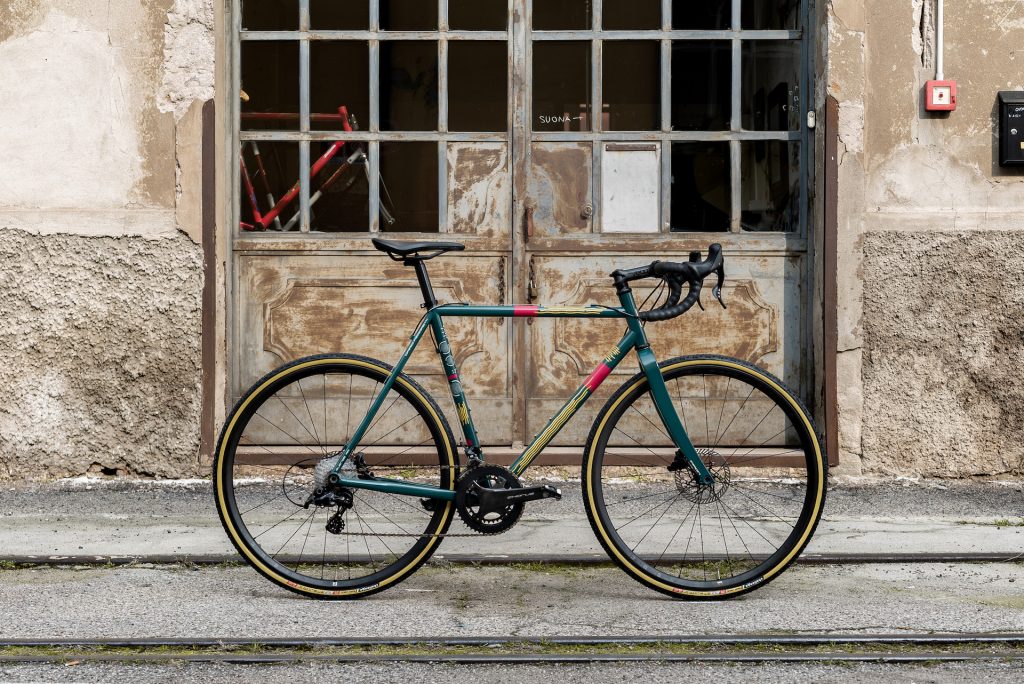 Pegoretti's Duende Rock & Roll, in front of the officina. The forest-green paint has yellow lines and red panels, and sits in front of a large, old garage door.