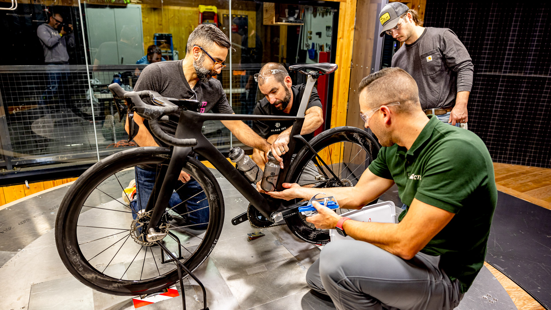 The image shows a group of engineers and a bike in the wind tunnel. The engineers are discussing something about the down tube bottle.