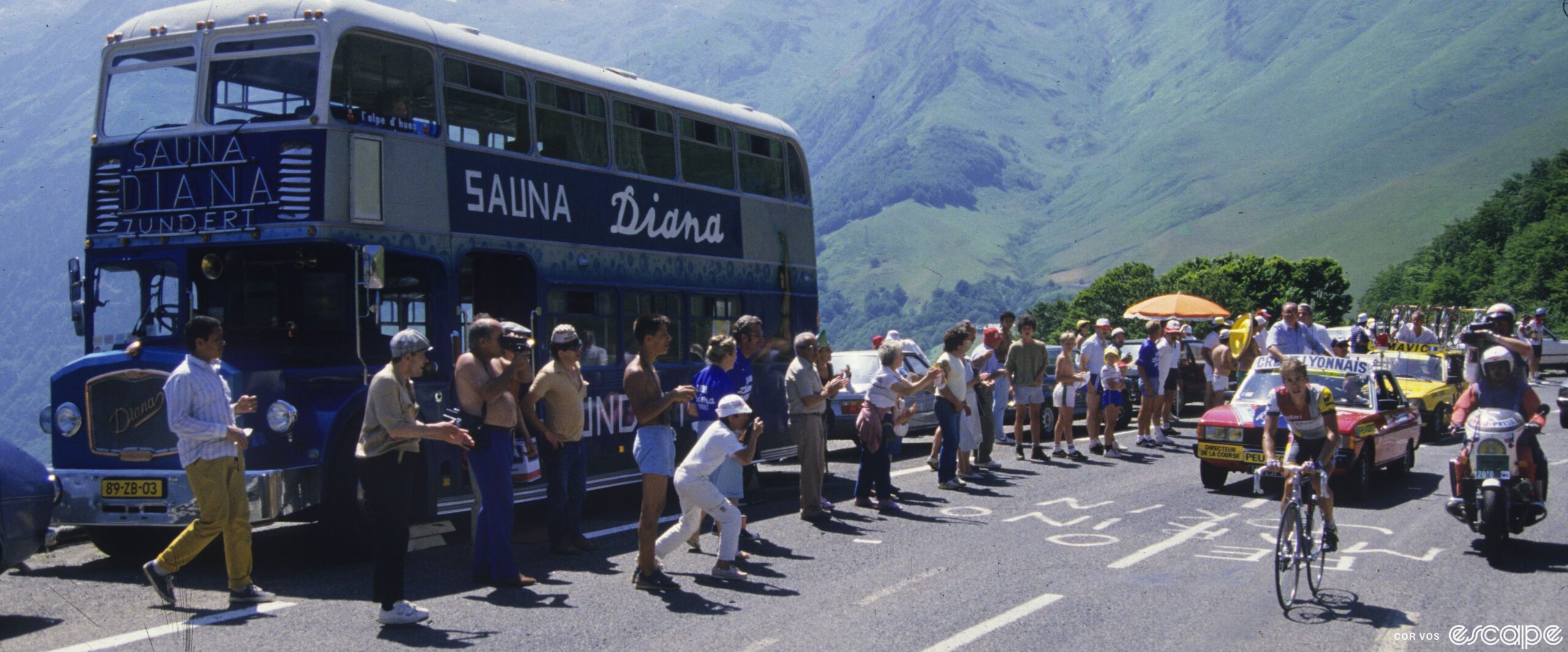 Andy Hampsten rides in the 1986 Tour de France. He's on a climb passing a double-decker bus labeled "Sauna Diana" and is followed by the red officials car and a yellow Mavic neutral support vehicle. Fans line the road to cheer.
