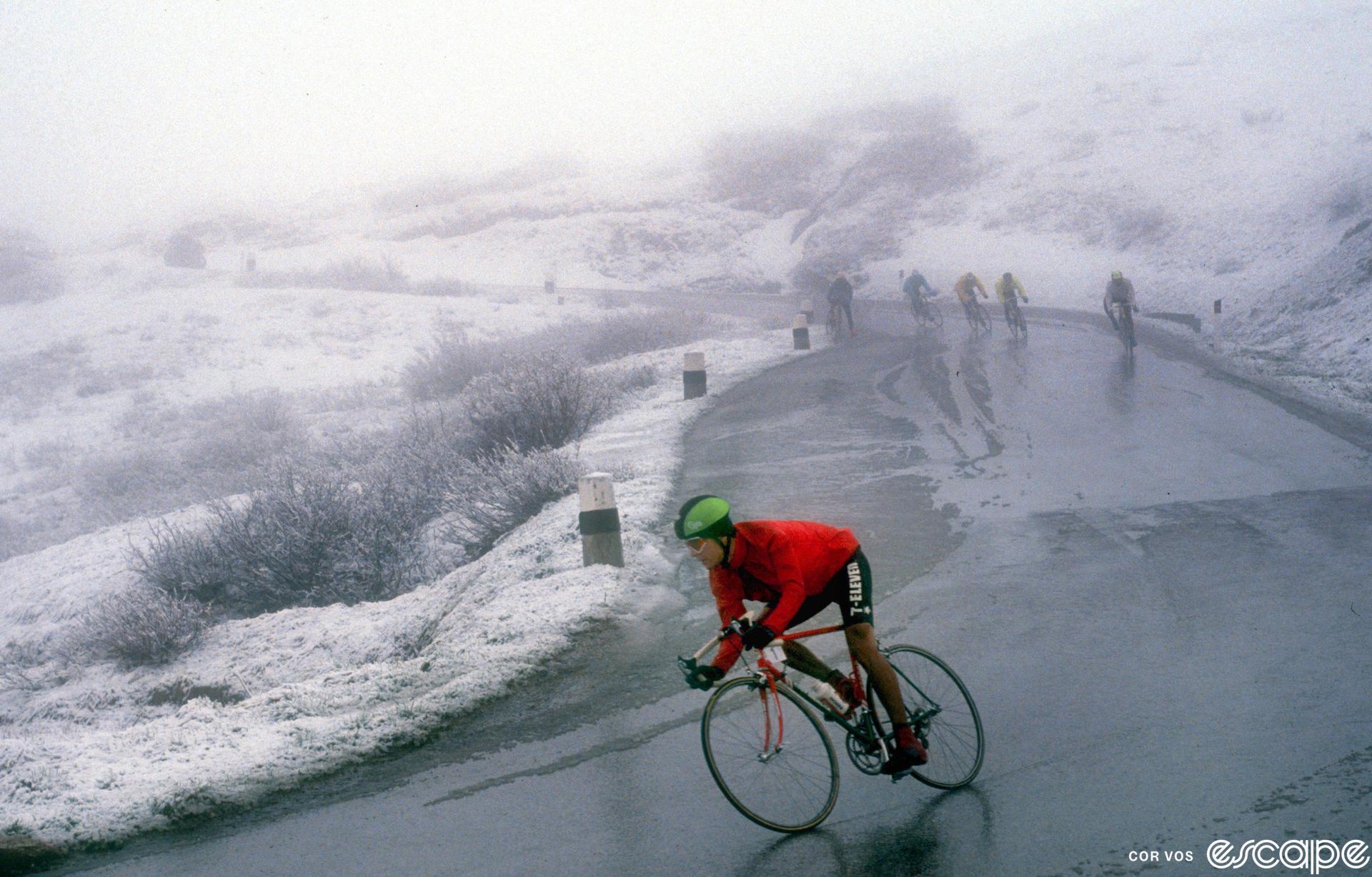 Andy Hampsten descends a fog-shrouded, snowy Passo di Gavia in the 1988 Giro d'Italia. He's wearing a fire engine-red jacket and full neoprene gloves as well as a helmet cover and skullcap, but his legs are bare. Behind, other riders descend on wet, freezing roads.