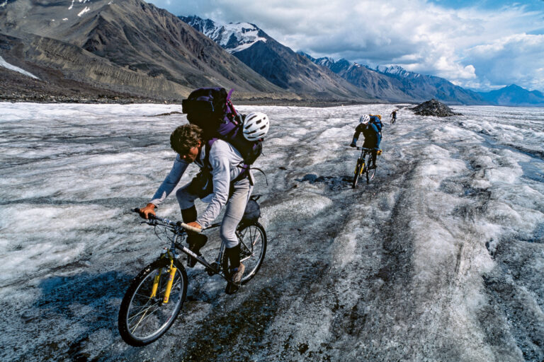 Three cyclists ride across a frozen glacier underneath towering snowy peaks. They're on older titanium hardtails, and as this was pre-bikepacking era, they're carrying all their gear in backpacking backpacks.