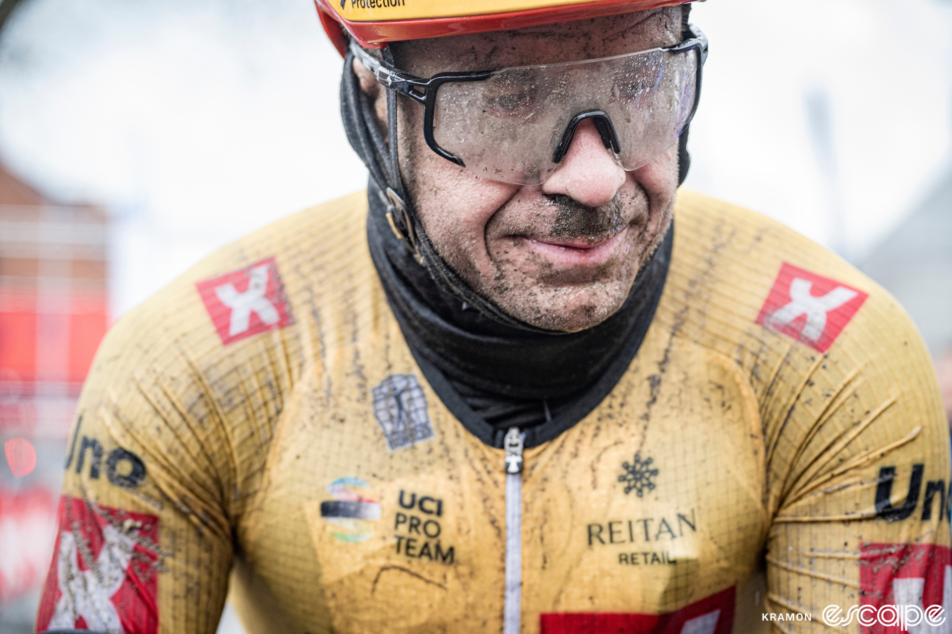 Alexander Kristoff sits at the finish line of the 2023 Gent-Wevelgem race. He is covered in grit, his glasses, face, and jersey speckled with grime and his jersey and balaclava soaked. He has a grim expression.