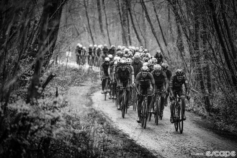 A pack of riders navigates the dirt (or mud in this case) Plugstreet section of the 2023 Gent-Wevelgem. The black and white image shows the riders in a pack, close together almost as if huddled for warmth, as tall trees and mist press in on both sides.