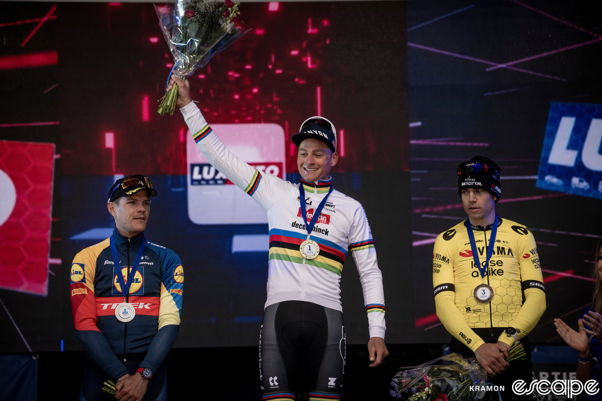 From left to right, Jasper Stuyven, Mathieu van der Poel and Wout van Aert stand on the podium at E3 Prijs. Van der Poel steps forward with a bouquet of flowers raised in one hand and a first place medal around his neck. To his left, Van Aert, with a third-place medal, looks on with a sideways glance and neutral expression.