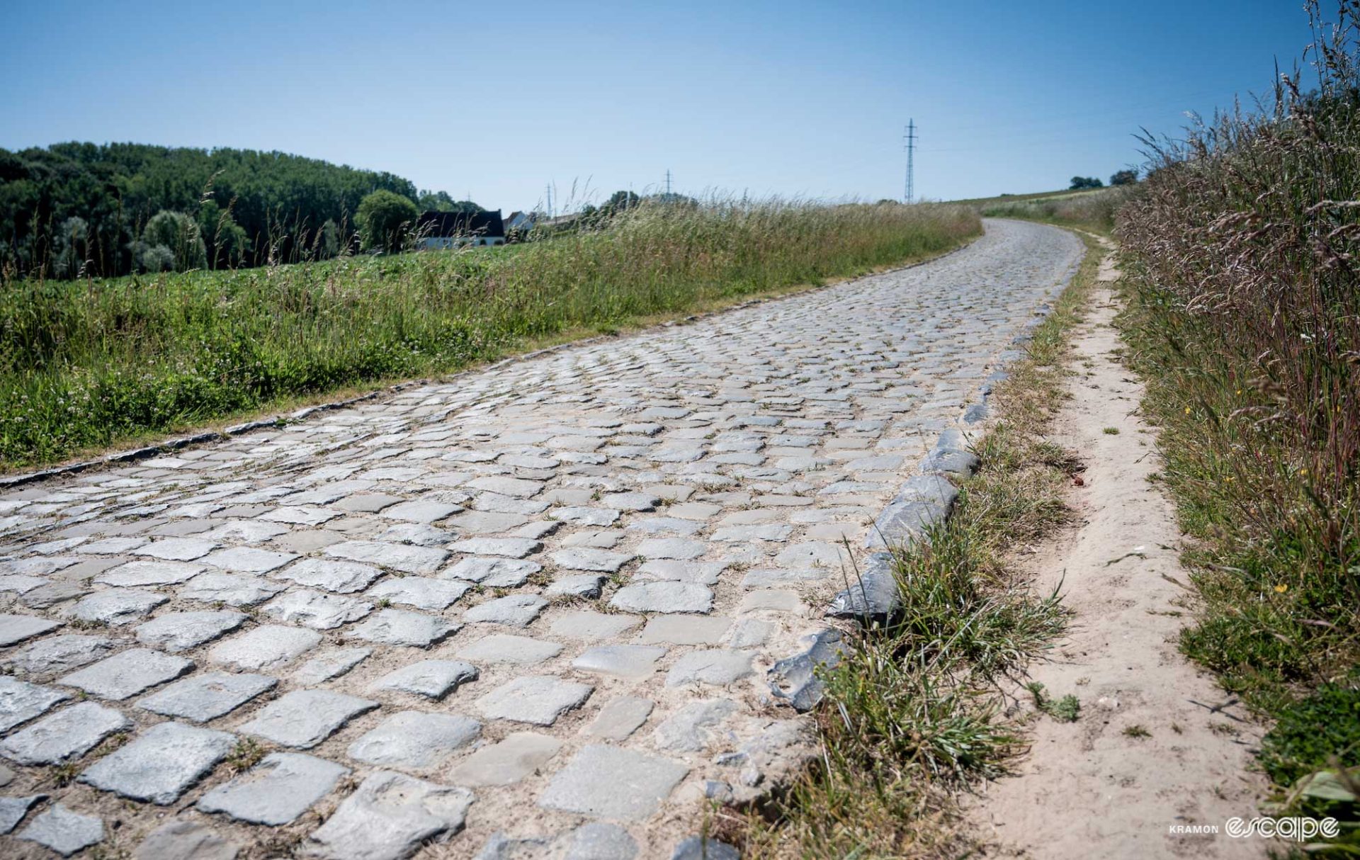 A blue granite cobbled lane rises through a field in the Flemish Ardennes. No one is in sight and it curves around a corner out of view.
