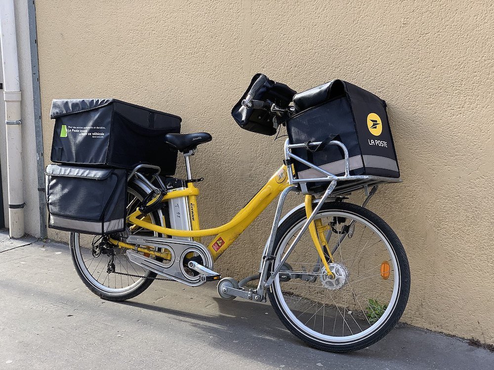 A yellow La Poste electric delivery bike, heavily laden with big black bags for letters. It is deeply utilitarian and unattractive. 