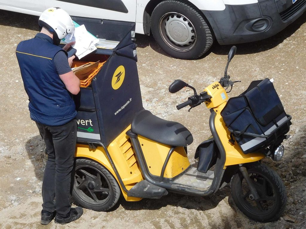 A three-wheeled yellow electric scooter with a big box on the back, a pouch on the front, and a man sorting letters for delivery standing next to it.