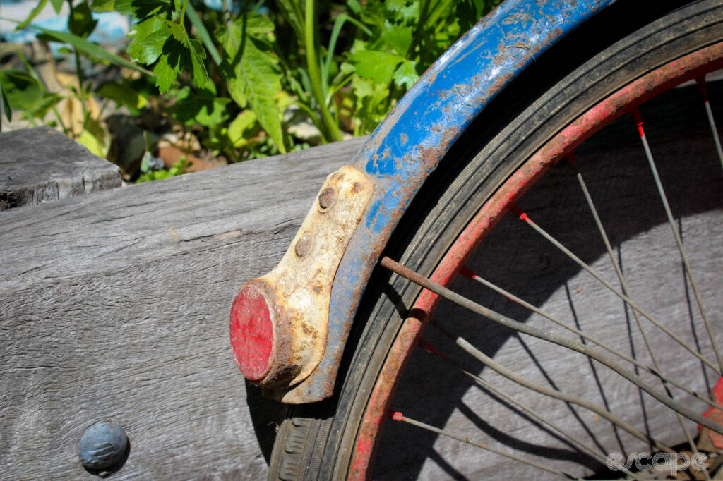 A rickety rear mudguard and rear wheel leans against a garden bed. 
