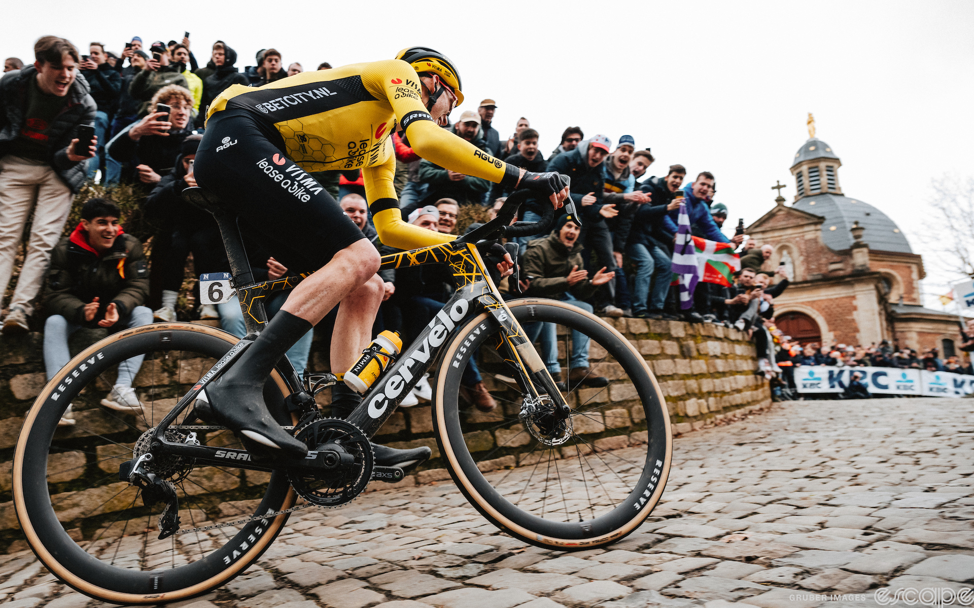 Matteo Jorgenson rides a solo breakaway up the Muur van Geraardsbergen at the 2024 Omloop Het Nieuwsblad. He's alone, at the top with the chapel looming on the right side of the picture. Fans lean over the stone wall to cheer. He's on an aero Cervelo S5 with 40/44 mm wheels, a skinsuit, and aero shoe covers.