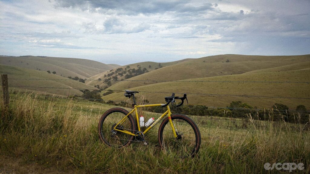 The author's bike, a yellow Ritchey Outback, leans against a barbed wire fence at the top of Sellicks Hill, overlooking huge rolling hills. 