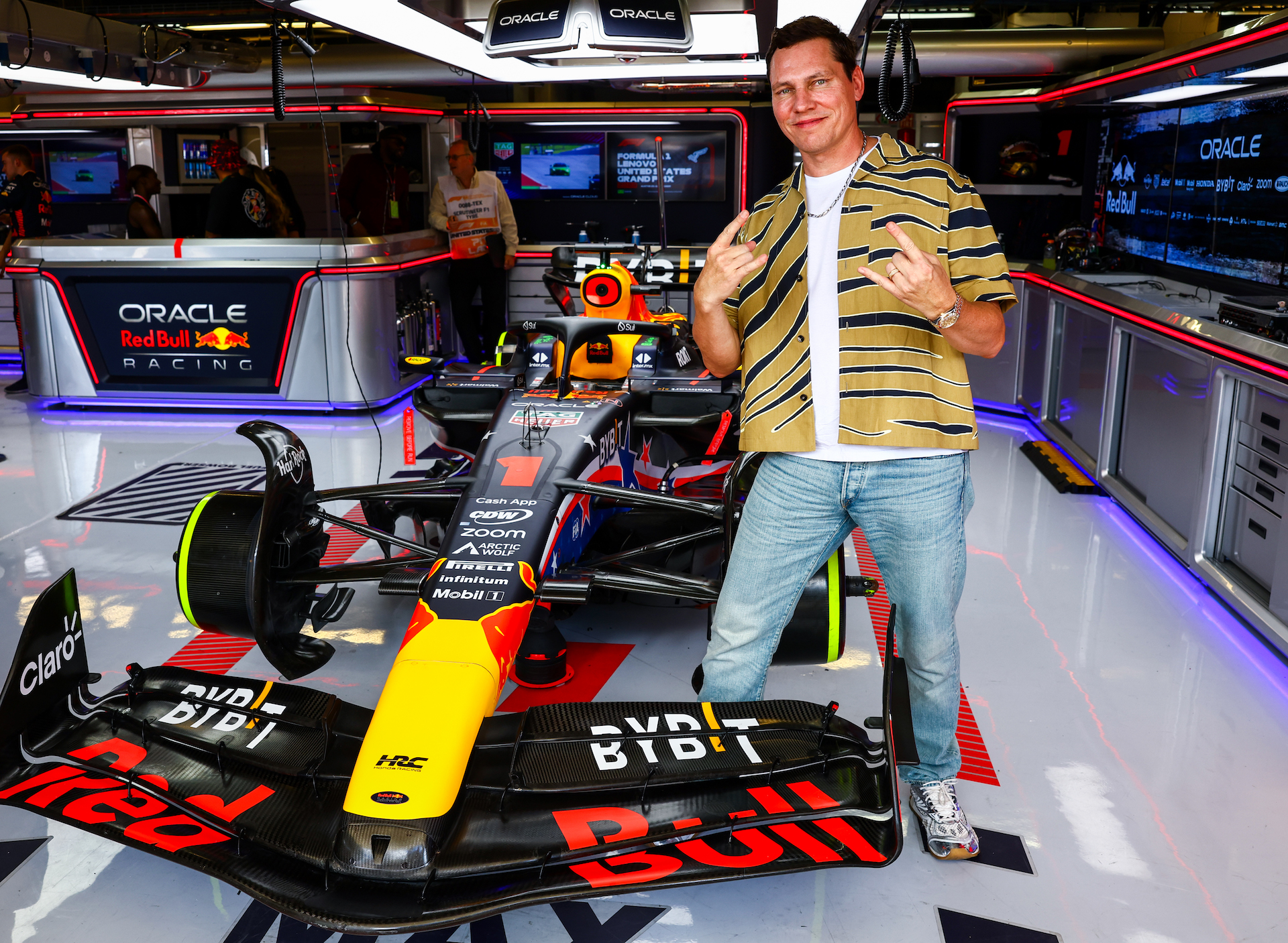 Dutch DJ Tiesto makes the "hook 'em horns" gesture with both hands as he stands in the Oracle race paddock at the Austin Grand Prix. He's standing over one of the race cars, quite close actually, in a way that only VIPs can get to a multi-million dollar precision race car. He is dressed casually in jeans and a tiger-striped short sleeve button down, open over a white t-shirt, and has a satisfied look on his face.