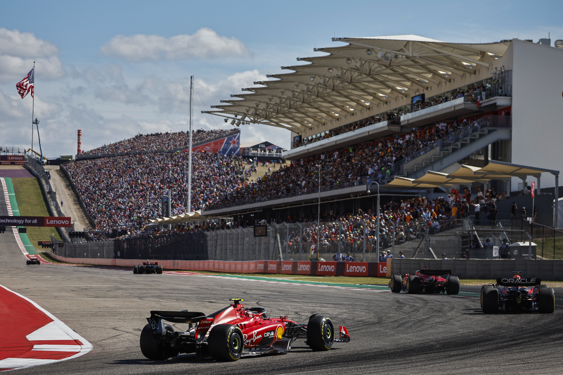 Charles Leclerc leads Max Verstappen and Carlos Sainz through a corner on the Austin circuit. They're about to pass a towering grandstand before driving through the start/finish and up a steep hill past another grandstand, also packed with fans.