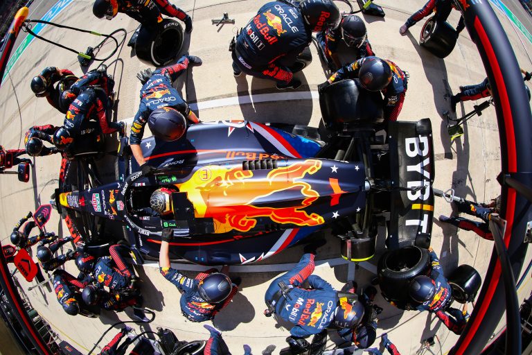 Max Verstappen's F1 car is surrounded by workers on pit row. The overhead shot captures the explosion of action as 22 workers swap all four tires in around two seconds.