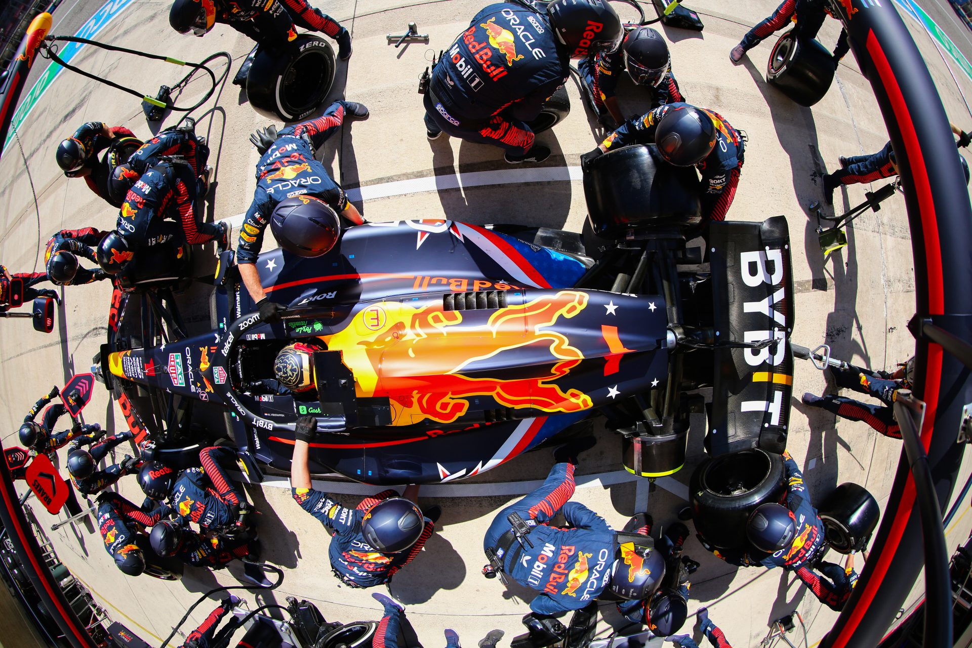 The Oracle pit crew attends to a race car in the pits. The fisheye overhead perspective distorts the view, capturing all 22 pit workers as they race to swap tires and fuel the vehicle, which sits at the core of the photo with its large red bull logo in the center.