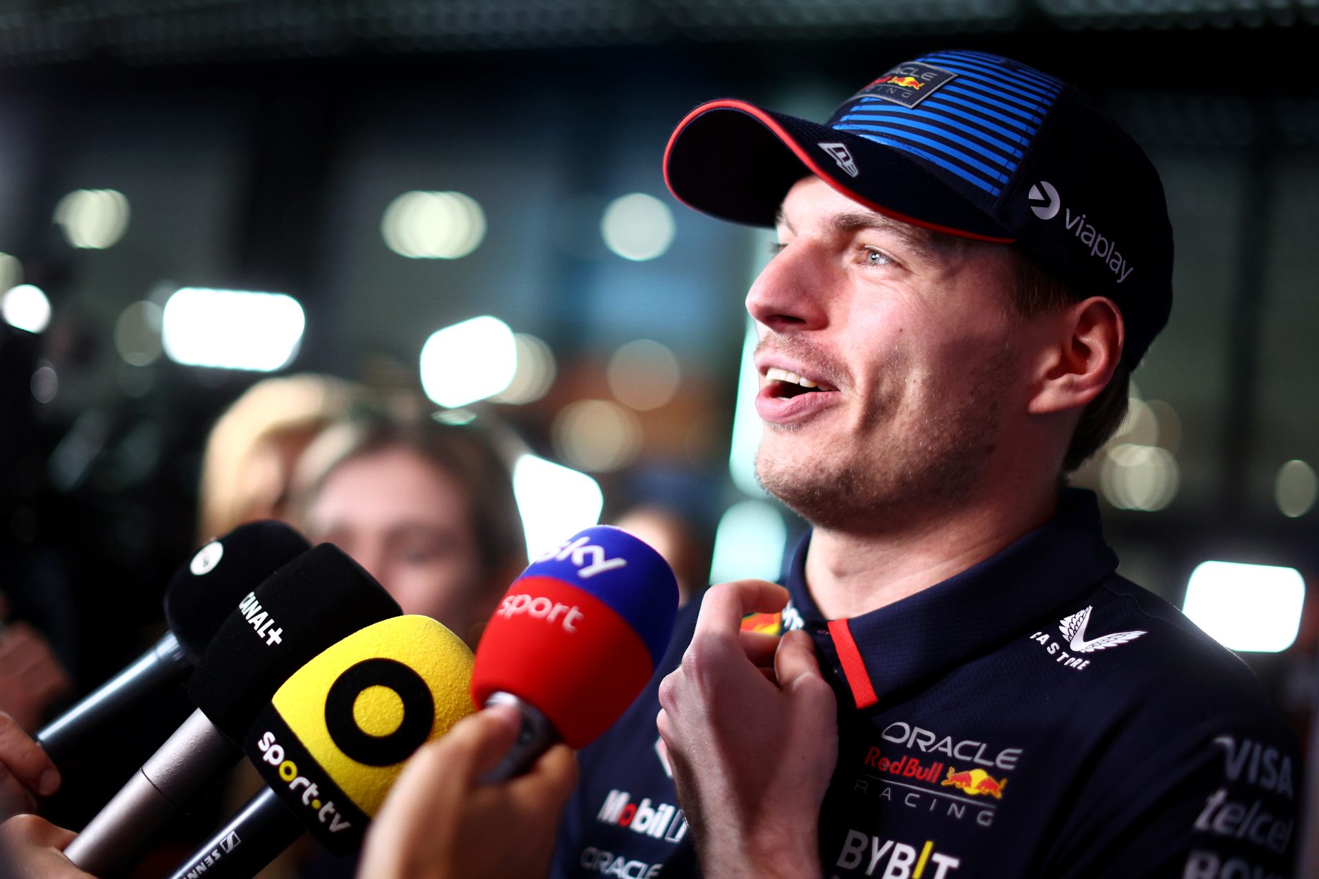 F1 star Max Verstappen speaks into a forest of media microphones in front of him. He has a smile and looks slightly above the heads of his unseen questioners and is clad head to toe in sponsor-logo gear.