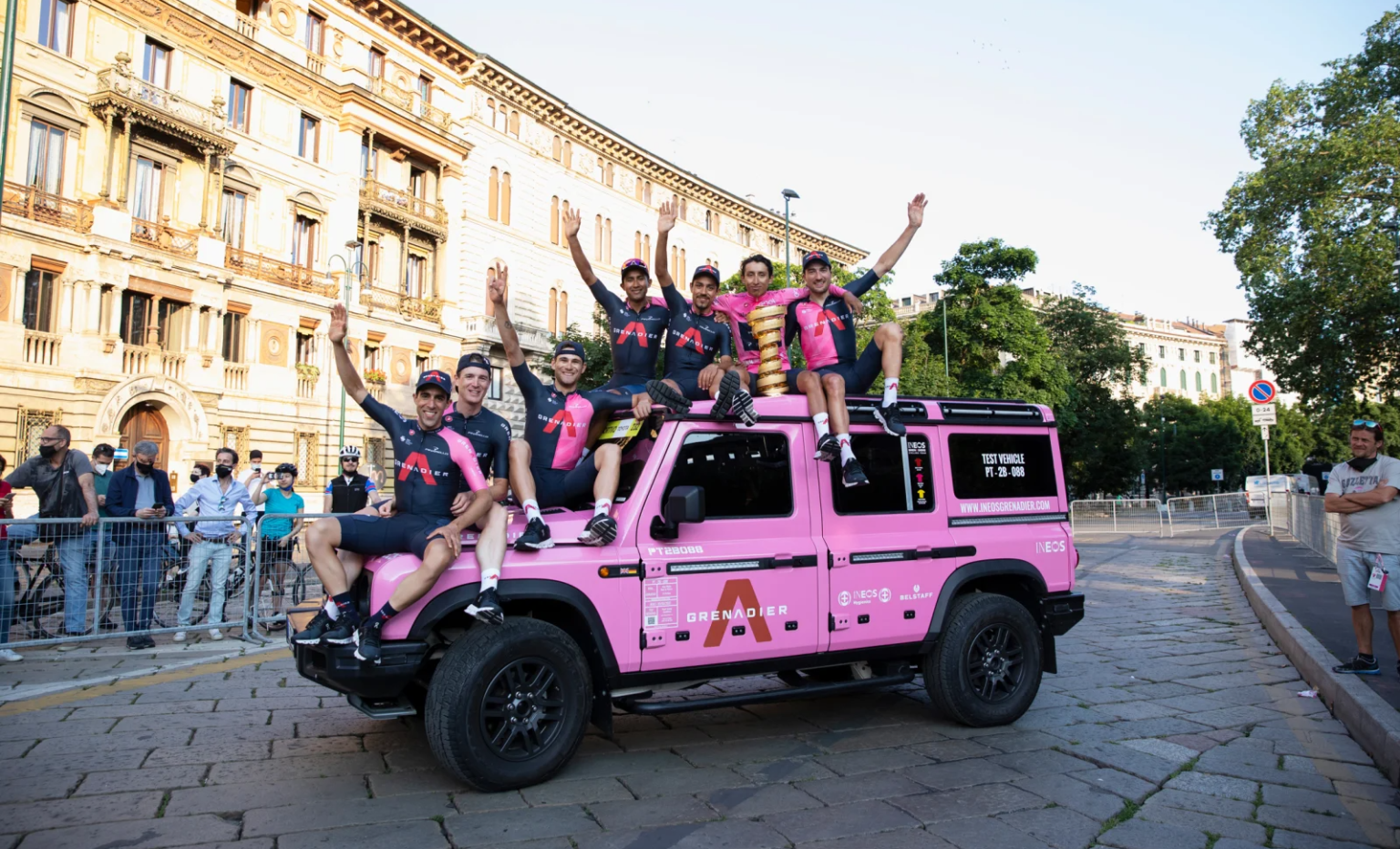 Seven Ineos Grenadiers cyclists sit atop a hot pink Ineos Grenadier vehicle in Rome. They are celebrating Egan Bernal's Giro d'Italia victory, and Bernal, in his matching pink jersey, sits in the center of the image atop the truck with the Giro's spiral-shaped trophy.