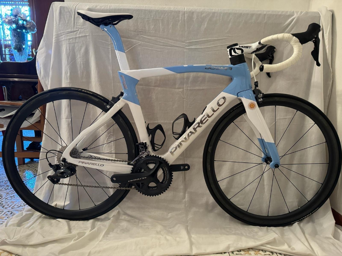 The Papal Pinarello, in light blue and white with custom details, sits on a crumpled white sheet against some furniture in the corner of a room somewhere. 
