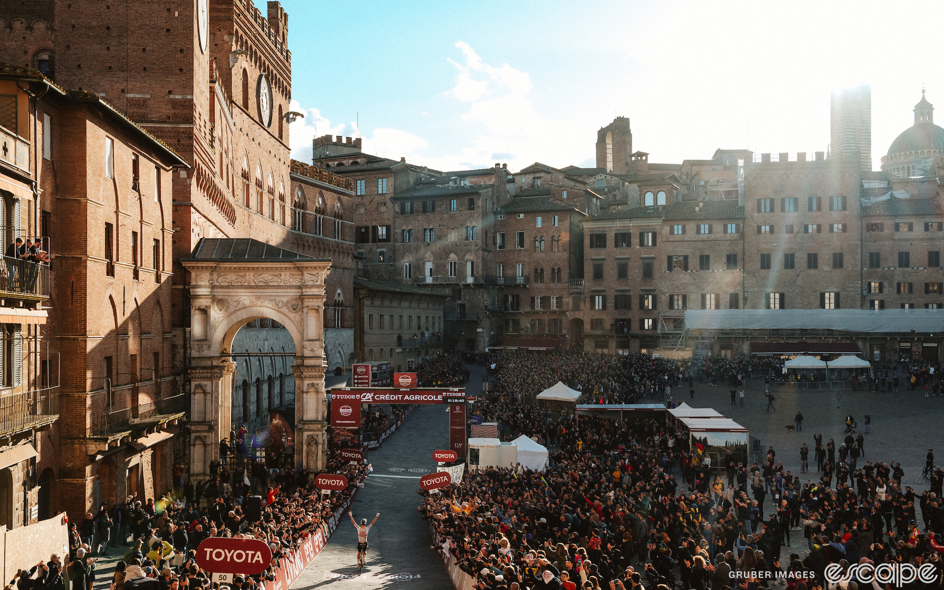 Tadej Pogačar raises both arms over his head as he crosses the finish line alone to win the 2024 Strade Bianche. He is shot from behind, sitting up as he approaches the finish gantry next to the arch and tall clocktower in Siena's Piazza del Campo. Large crowds gather in the square, and the setting sun lights up Pogačar and the building next to him.
