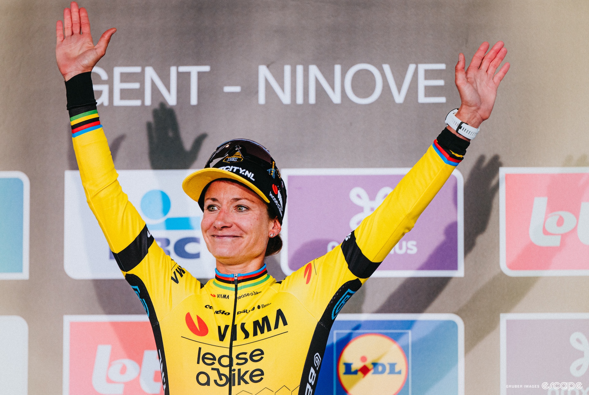 A woman in cycling clothing raises her arms over her head on a podium 