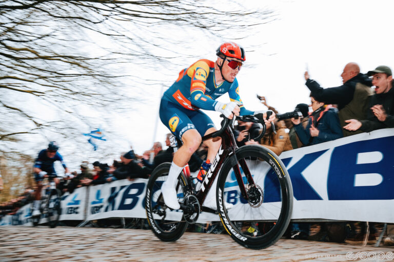 Mads Pedersen races up a hellingen in the Dwars door Vlaanderen. He's shown in a blur of speed, his primary-color kit bleeding into the pale white sky above and behind him.