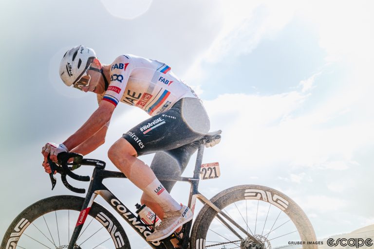 Tadej Pogačar dips his head from the effort of his 80 km solo breakaway at the 2024 Strade Bianche. He's slightly backlit, the soft glow of a flash illuminating him against a dreamy pale blue sky with billowing white clouds. His bike and kit are filmed in a white coat from the gravel roads.