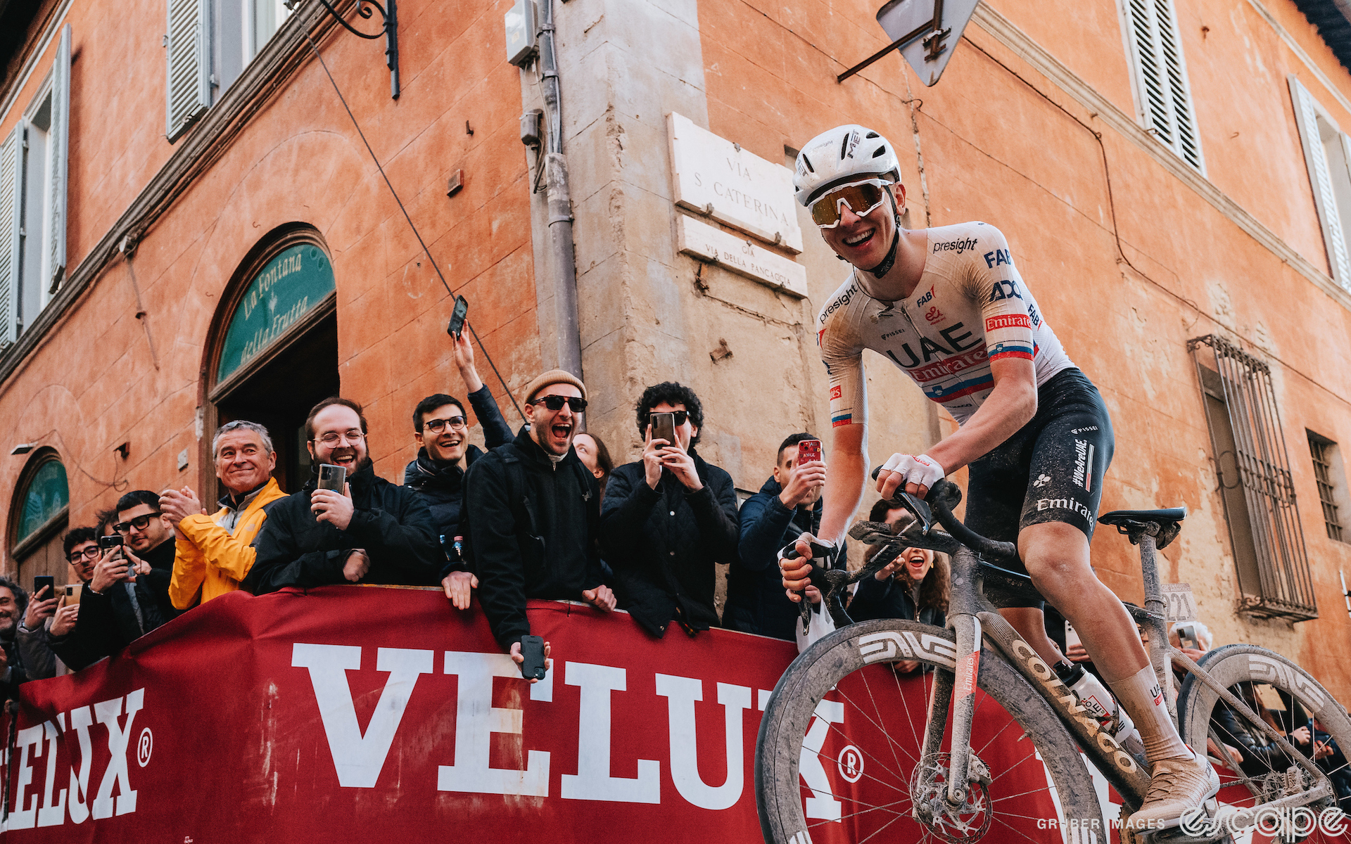 Tadej Pogačar crests the top of the Via Santa Caterina climb into Siena at the 2024 Strade Bianche. He is mugging for the camera, facing the photographer and looking down at the lens with a wide, ebullient grin. Behind him, fans respond in kind, with big smiles and cheers as they applaud.