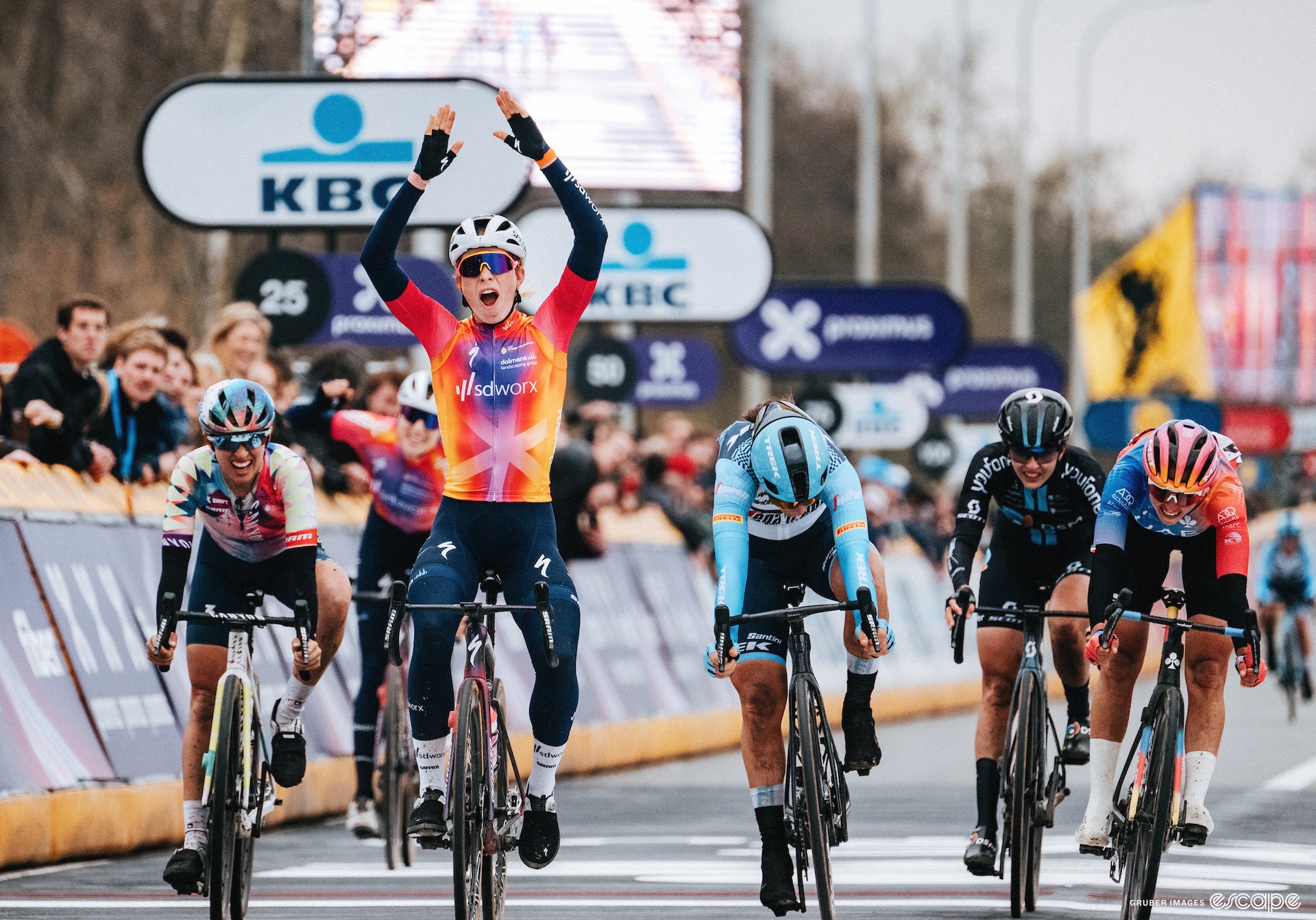 A cyclist raises their arms in the air at the finish like of a race. She is surrounded by other riders who look like they are in pain. 