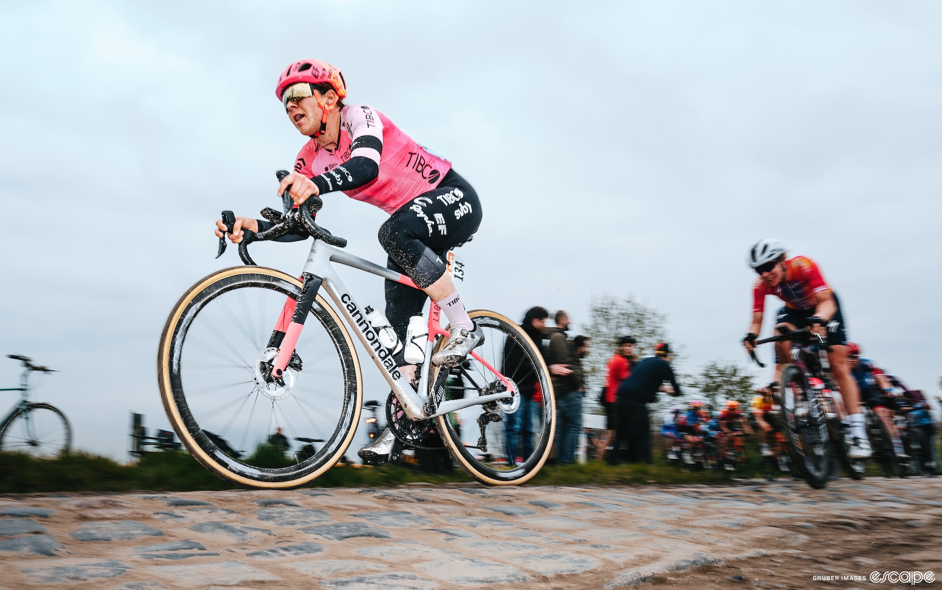 Jackson floats over the cobbles. She's shot from slightly below, looking up at her, and there's a blur to the shot. The flash illuminates her bright pink jersey against the pale white cloudy sky.