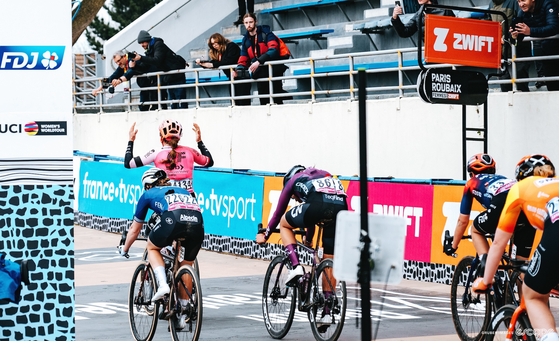 Alison Jackson sits up as she wins Paris-Roubaix Femmes. She shown from behind, at the front of a small group of riders, and has her hands in the air as she crosses under the finish gantry.