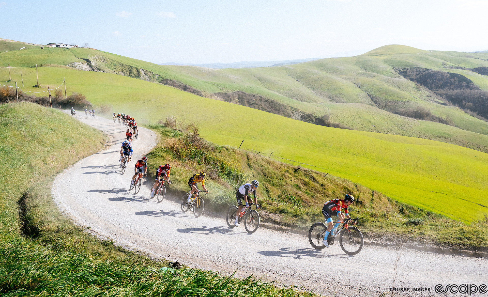 Riders negotiate a corner on one of Strade Bianche's gravel sectors. Dust rises from the line of riders as they sweep through turns amid green grassy fields and low hills.