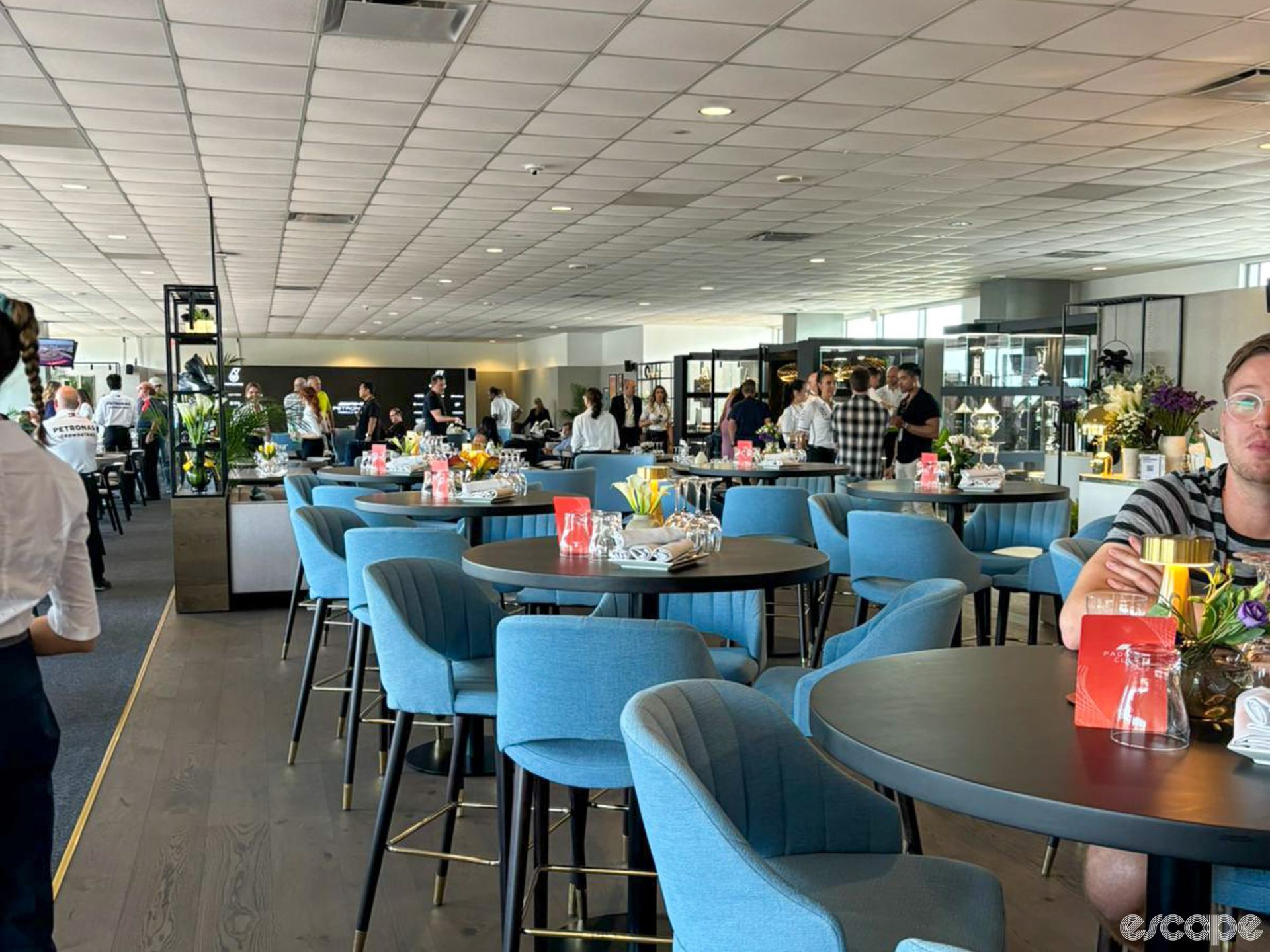 A sparsely populated VIP area at the Austin Circuit of the Americas course. Modish blue chairs sit at hightop tables, each with a careful centerpiece of flowers, napkins and glasses. Behind, small clusters of racegoers talk.