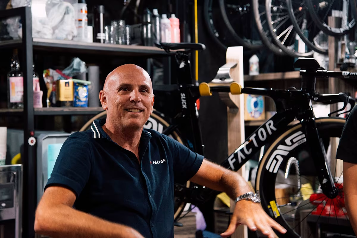 Rob Gitelis, CEO of Factor Bikes, sits at the brand's offices in a shop room. behind him, one of Factor's Slick triathlon bikes sits on a display rack in front of shelving for shop supplies.