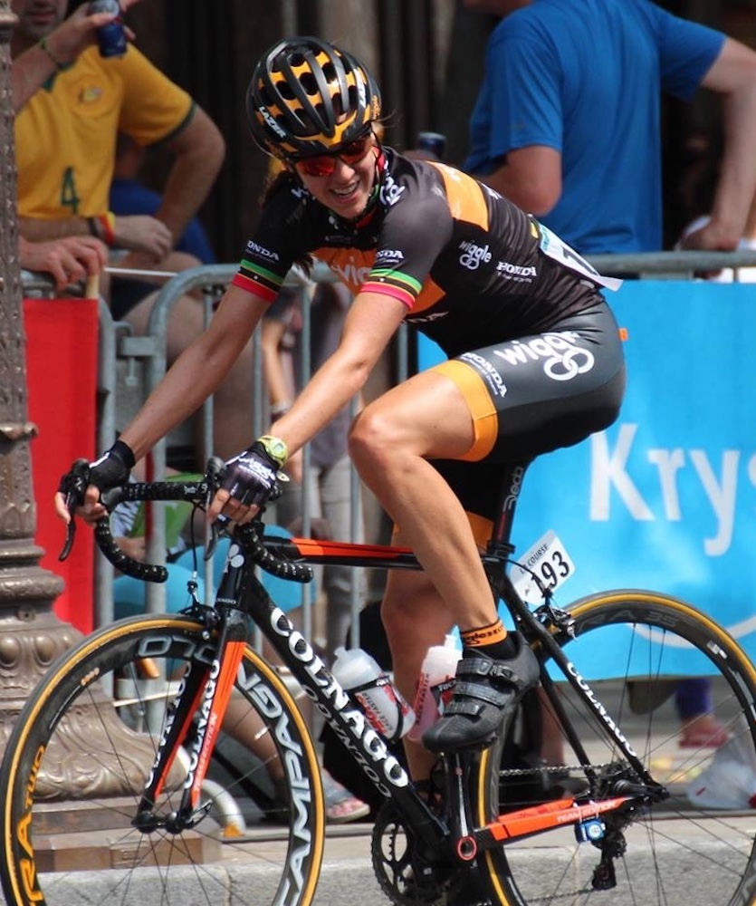 Kathryn Bertine rides in the 2014 La Course by Tour de France. She's dressed in the Wiggle team kit and aboard a Colnago as she looks over her left shoulder.