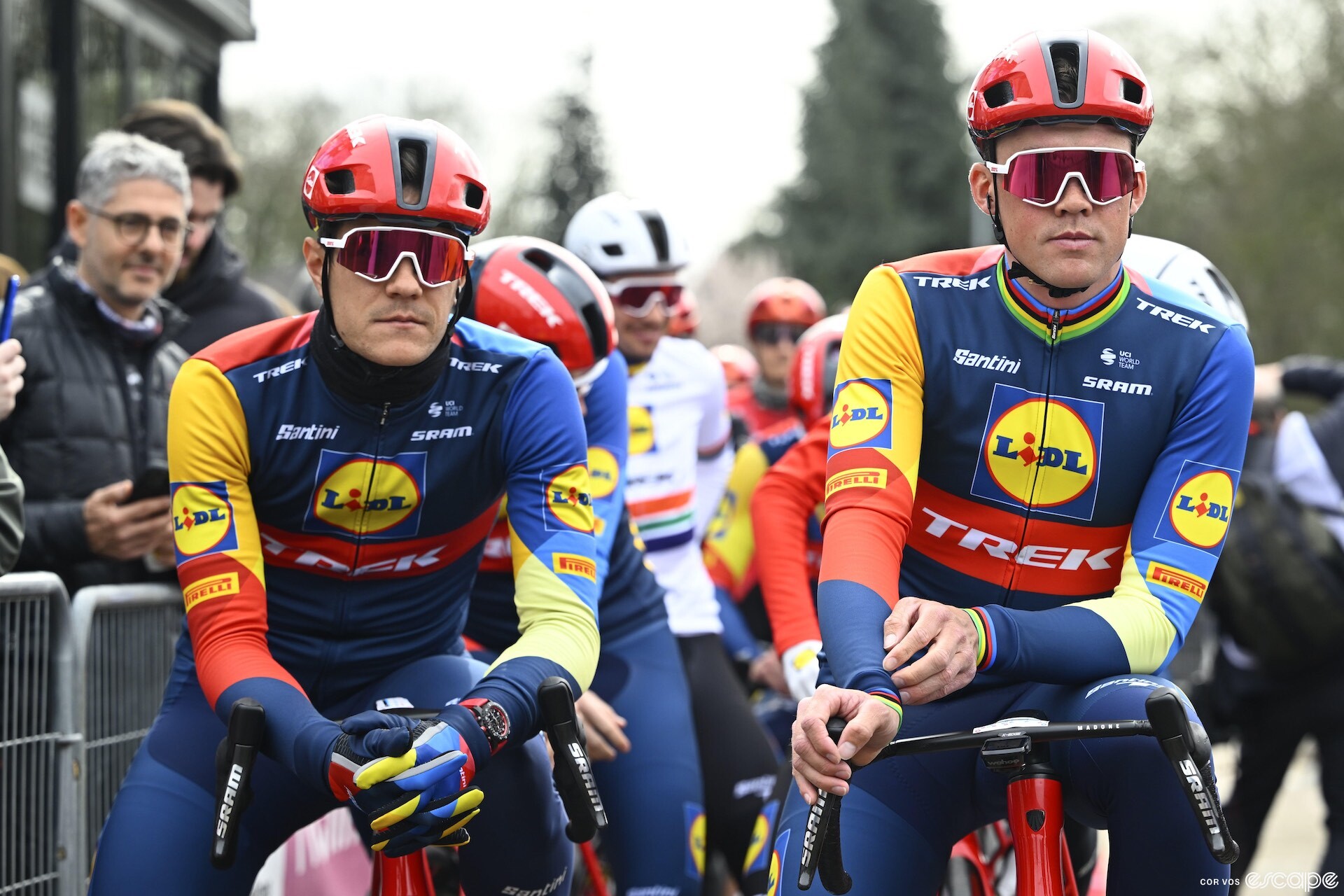 Jasper Stuyven and Mads Pedersen sit impassively at the start of a stage of Paris-Nice. Their eyes are hiddenn behind large sunglasses, and both are dressed warmly against a cloudy, cold sky.