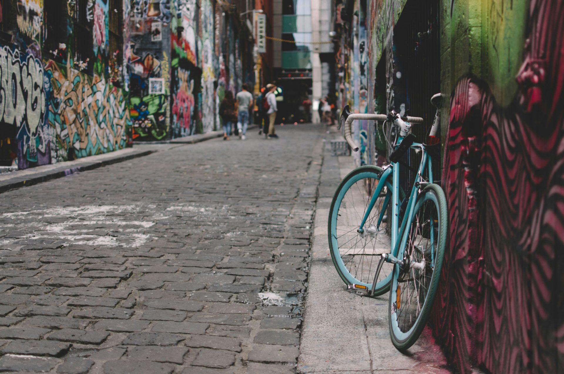 A light blue bicycle leans against a graffito'd wall in an alley in Melbourne. The alley is paved with bluestone cobbles, and the walls are covered in more brightly colored graffiti