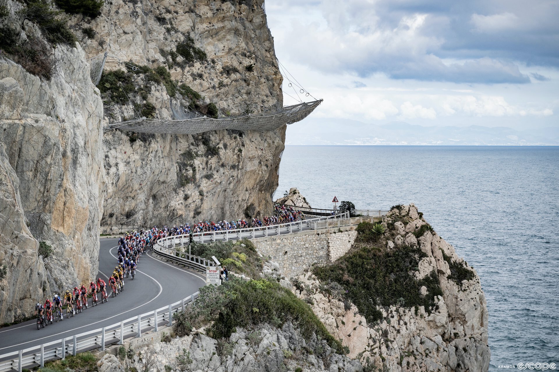 The Milan-San Remo peloton descends to the Mediterranean coast on a winding road, set into a sheer cliffside. Behind, the blue waters stretch out calmly under a partly cloudy sky, while in the distance the curving Italian coastline can just be seen.