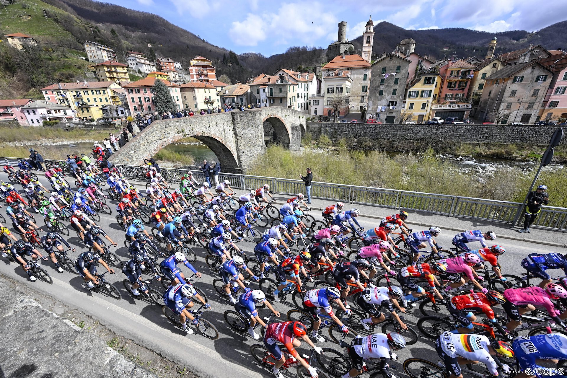 The Milan-San Remo pack passes a picturesque Italian village with red tile roofs. Fans are gathered on a pedestrian bridge over a stream to watch as the pack passes.