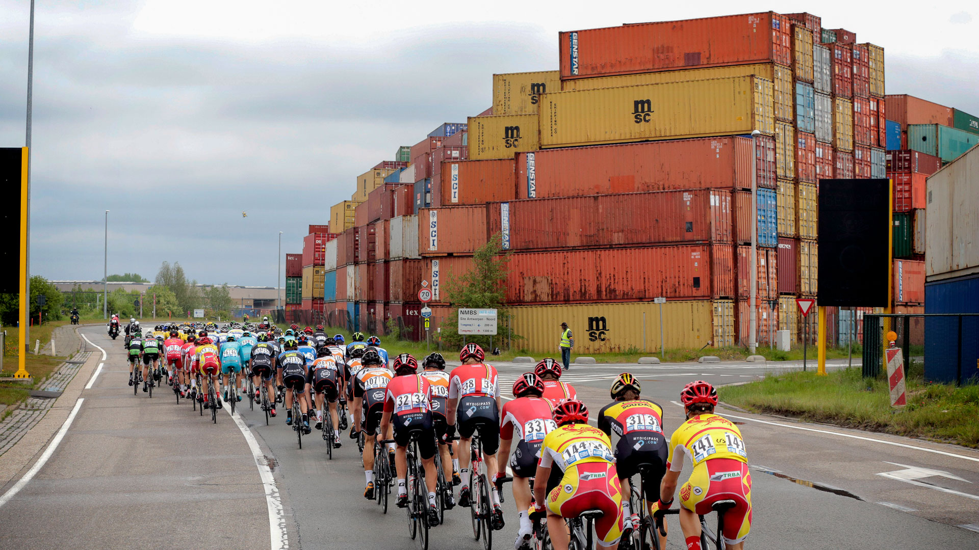 The image shows a bike race passing a tower of shipping containers.