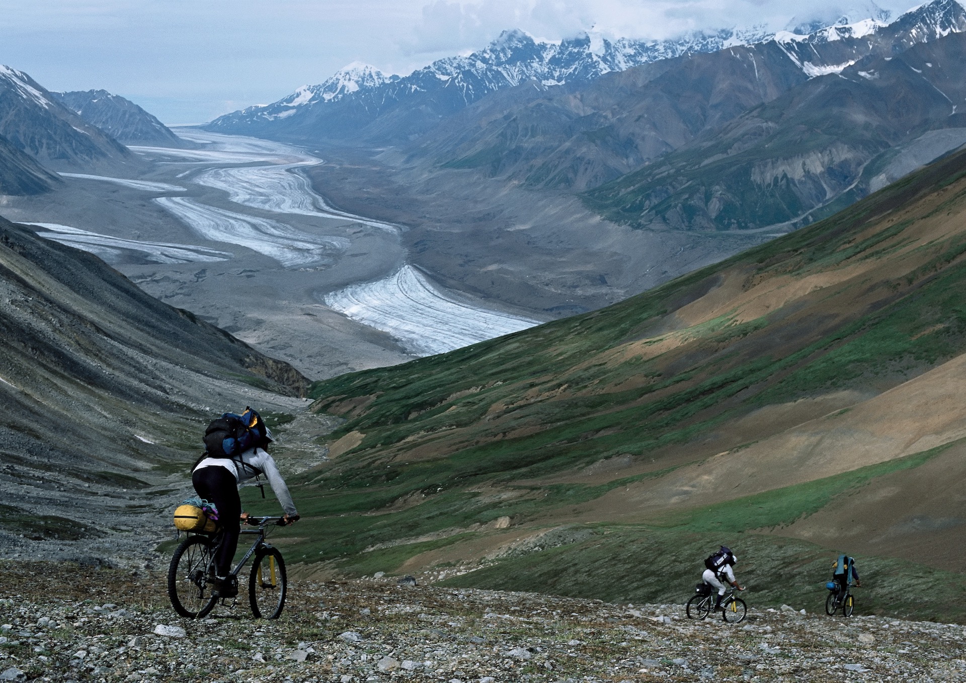 Three mountain bikers ride down a trailless scree slope above a striated glacier flowing through a huge, broad mountain valley.