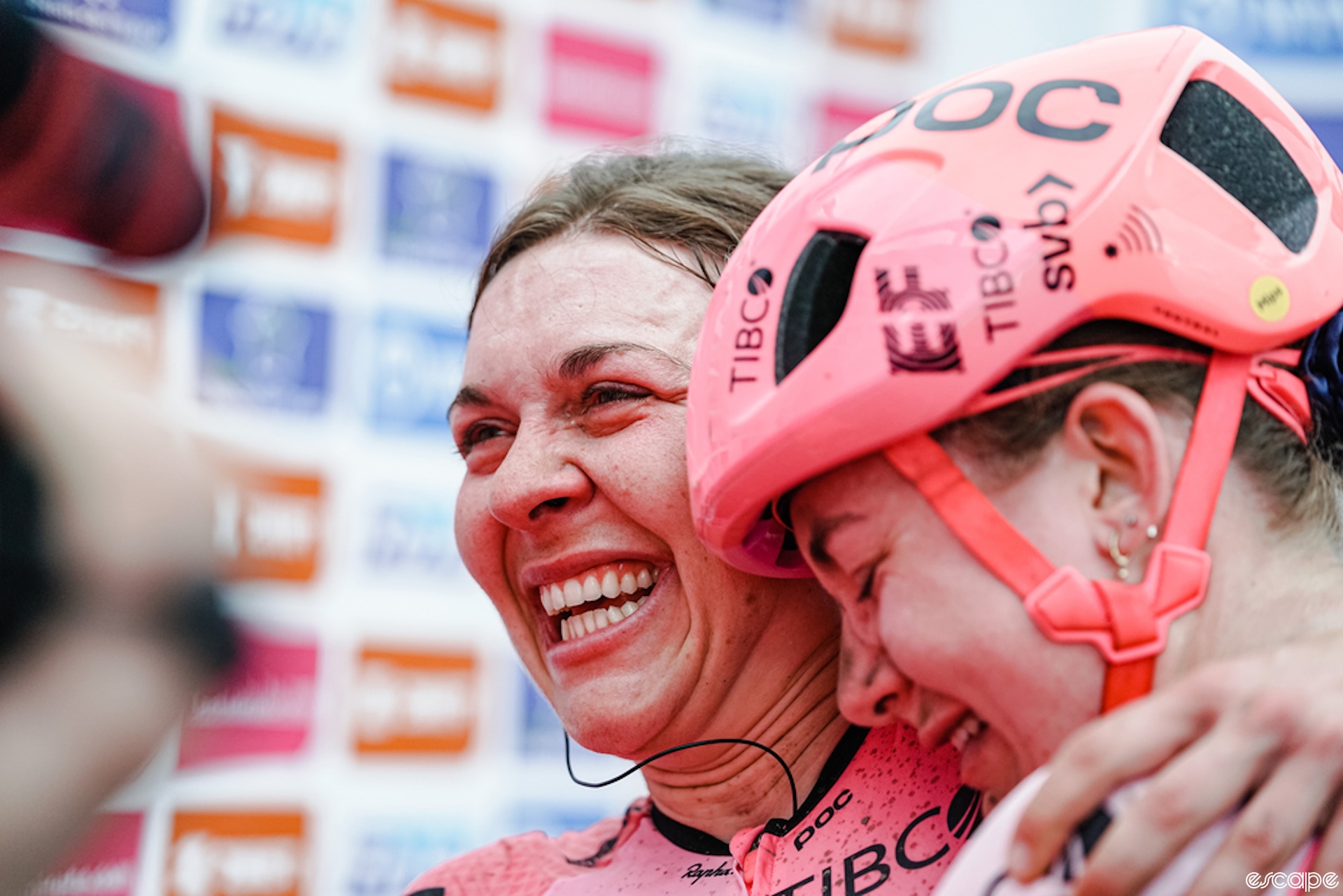 Alison Jackon smiles wide as she hugs a teammate after winning Paris-Roubaix. Her teammate has her eyes closed with deep emotion on her face as they embrace.