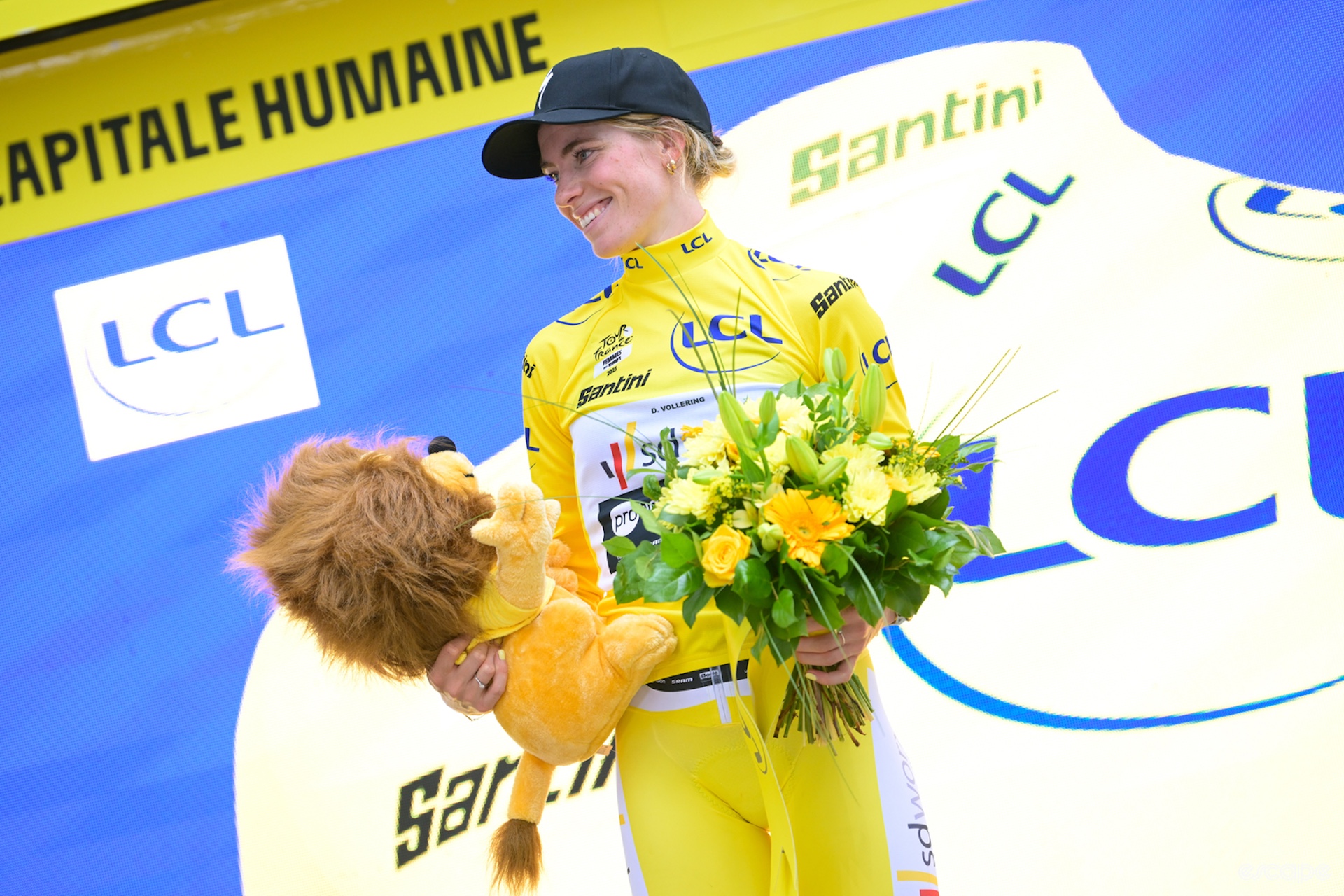 Demi Vollering, dressed in the yellow leaders jersey of the Tour de France, holds a lion and flowers and smiles at someone off picture to her right.
