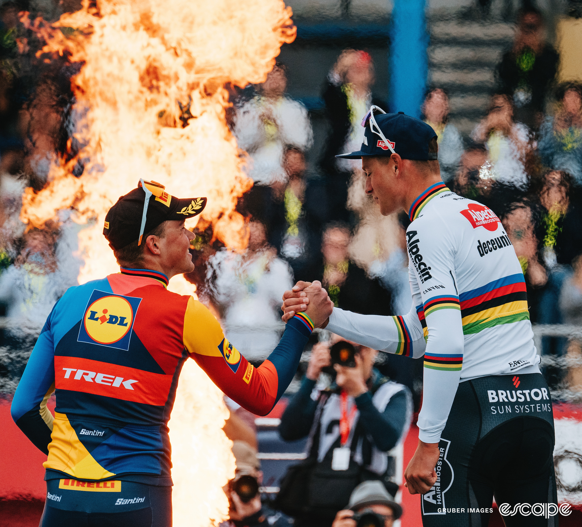 Mathieu van der Poel shakes hands with Mads Pedersen on the Paris-Roubaix podium. In front of them, flames spark from a pipe in the base of the stage and the crowd behind them shimmers in the heat-distorted air.