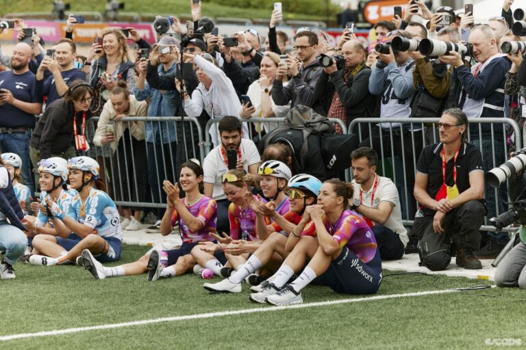 A cycling team of women sit on the ground while they watch their teammate hoist a trophy.