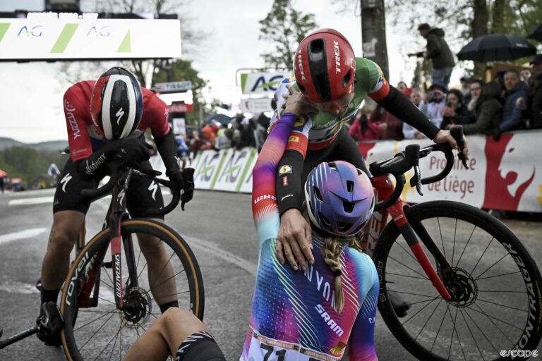Three women catch their breath at the top of a steep climb after a bike race, two are hugging