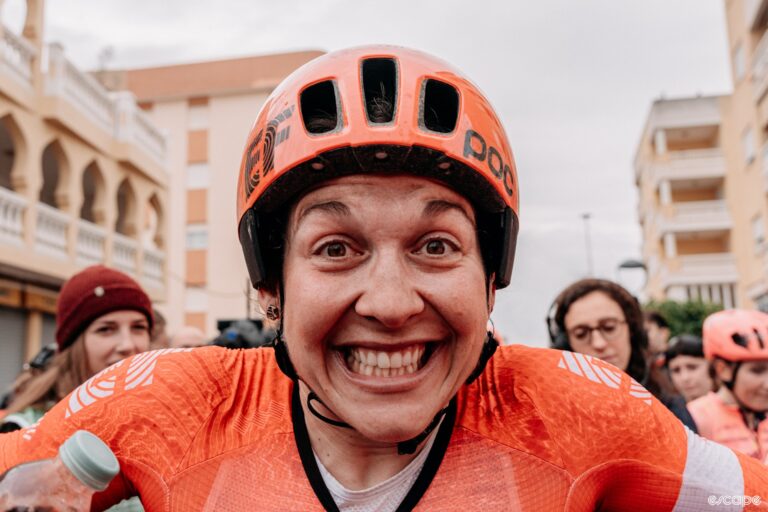 A woman in cycling clothing and with a dirty face gives the camera a huge smile.
