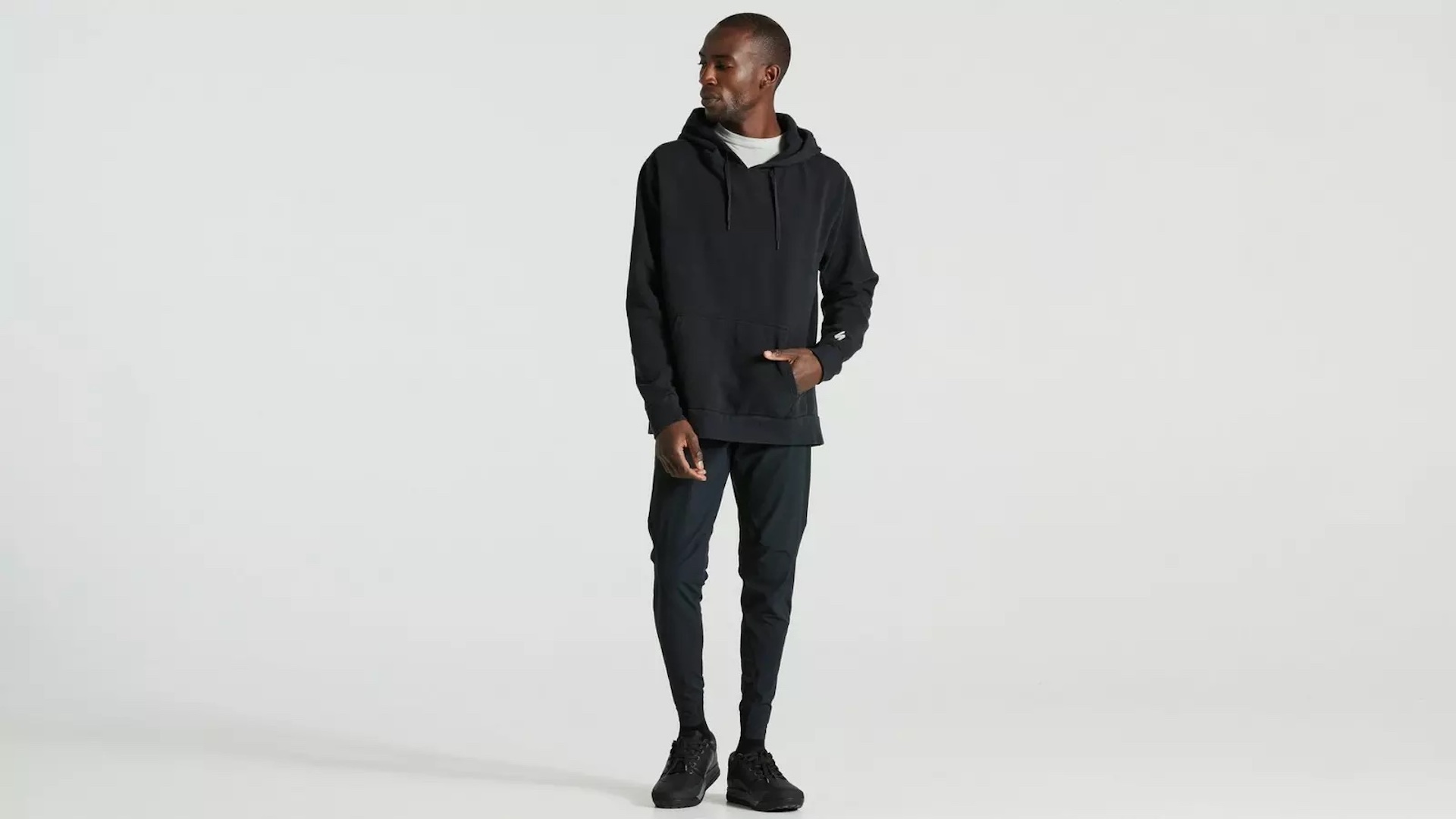 A black man models a Specialized cotton hoodie. He's dressed head to toe in black except for a white t-shirt and is looking contemplatively off his right shoulder.