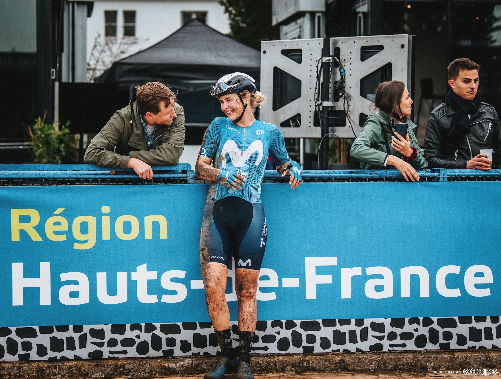 Emma Norsgaard leans against a fence at the finish of Paris-Roubaix; she is covered in mud, talking to a friend in normal street clothing. They both laugh. 