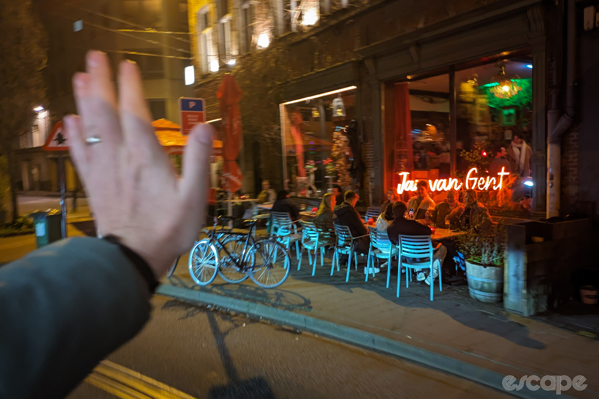 A hand waves in the foreground. A group sitting outside a bar do not reciprocate. 