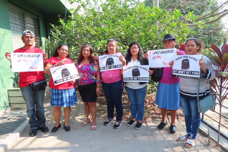 Seven Salvadoran workers, six of them women, stand in front of a camera holding signs that read in English and Spanish "Specialized did not pay me" and "I sewed your clothes but didn't get paid."